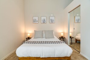 Un llit ben fet amb edredó blanc i almoades amb ratlles, flanquejat per taules de nit amb làmpades, en una habitació amb un mirall gran i tres obres florals emmarcades a la paret.