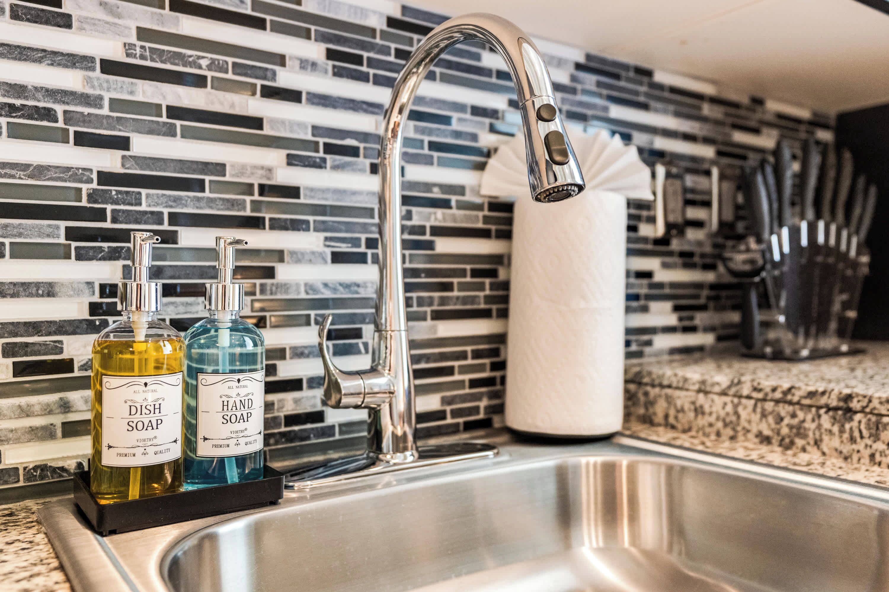 A kitchen sink with a modern faucet, two bottles of dish soap, and a paper towel holder.