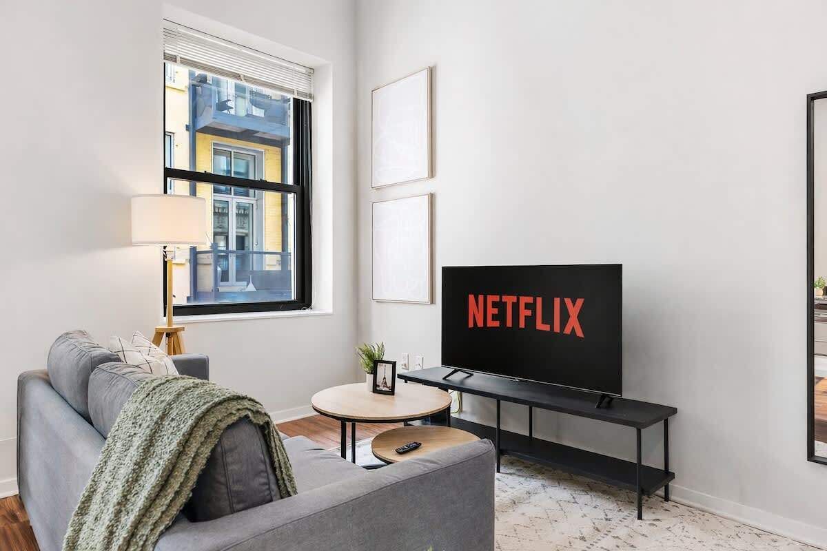 A modern living room with a grey sofa, a round coffee table, a black TV stand with a Netflix logo, and a window with blinds.
