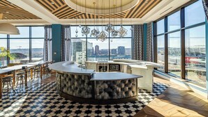 Eine moderne Bar mit einer zentralen Theke, großen Fenstern und Stadtblick sowie einem gemusterten Boden.