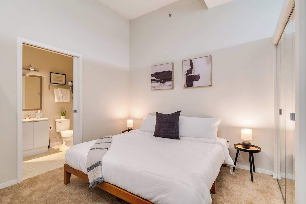 Une chambre à coucher avec un lit, des tables de chevet, une commode, une œuvre d’art accrochée au mur et une salle de bain visible à travers une porte ouverte.