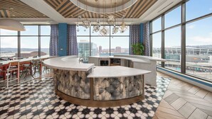 Eine moderne Hotelhalle mit einer zentralen Bar, großen Fenstern mit Blick auf die Stadt und einem gemusterten Boden.