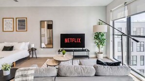 Et moderne hotelværelse med en stor seng, en sofa, et sofabord, et fjernsyn der viser 'NETFLIX', et spejl og et vindue med udsigt til bygninger.