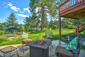 Un patio exterior con una chimenea, asientos y una terraza de madera con vistas a un jardín con árboles y una valla.