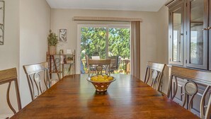 Ein Esszimmer mit einem langen Holztisch, Stühlen und einer Obstschale. Ein Fenster bietet Blick auf Bäume, und eine Tür führt in einen Außenbereich.