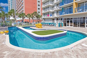 A swimming pool with a grassy area, lounge chairs, and a view of a tall building. - Atlantica 656 (Myrtle Beach)