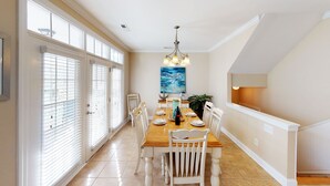 A dining area with a wooden table, white chairs, a chandelier, and a painting on the wall. - Caribbean Cove 504 (Myrtle Beach)