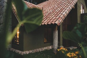 Une maison avec un toit en tuiles, un pilier en pierre et une fenêtre d’où s’échappe une lumière chaleureuse.