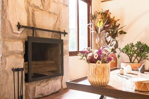 Un mur en pierre avec une cheminée, un vase en bois rempli de fleurs et une fenêtre donnant sur un paysage verdoyant.
