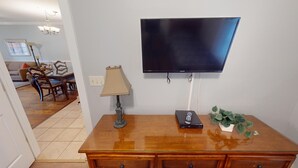 Ein Wohnzimmer mit Flachbildfernseher an der Wand, einem Holztisch, einer Lampe und einer Pflanze.