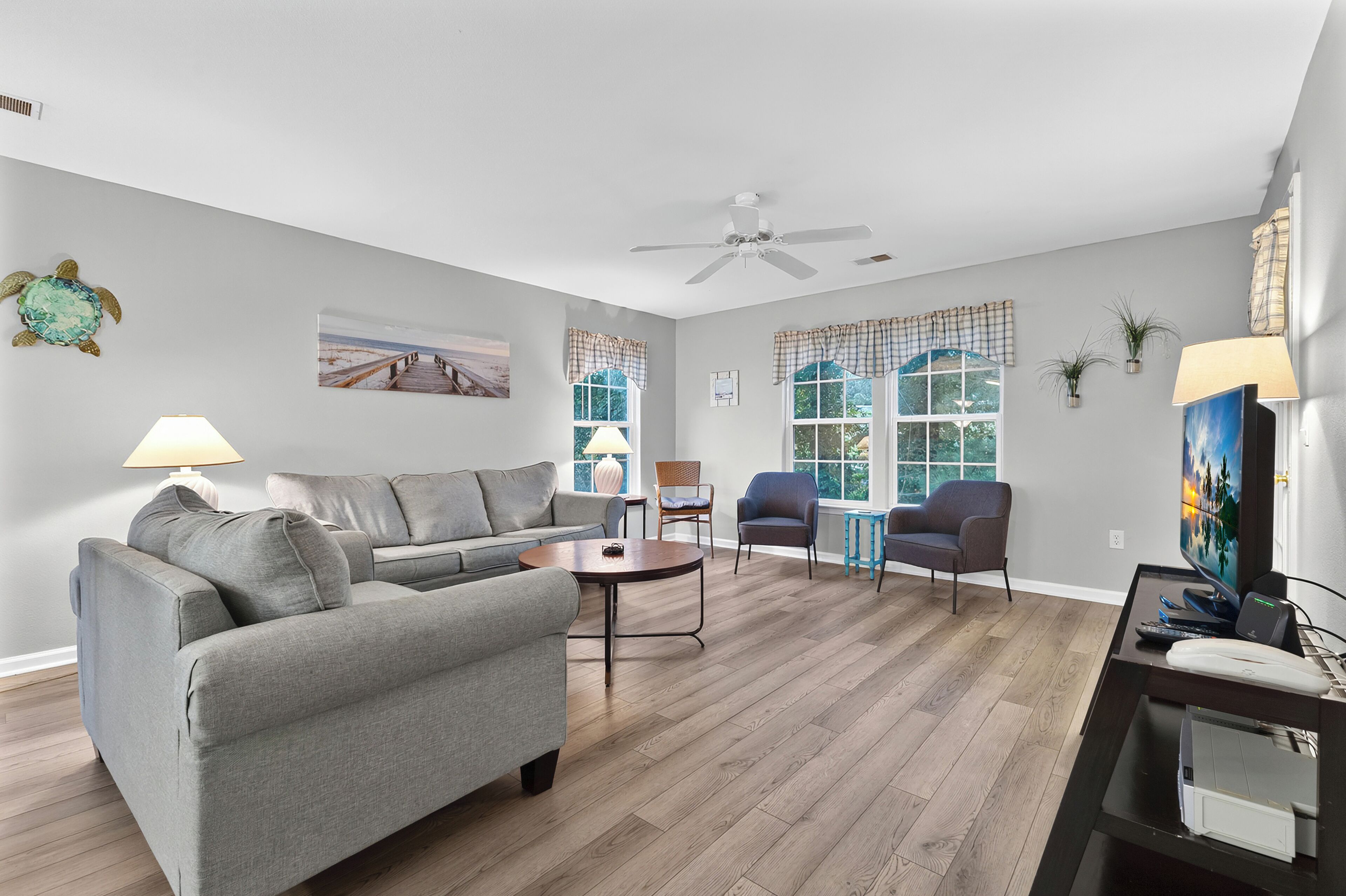 A living room with a grey sofa, a coffee table, and two armchairs. There is a ceiling fan, a TV, and a window with curtains.