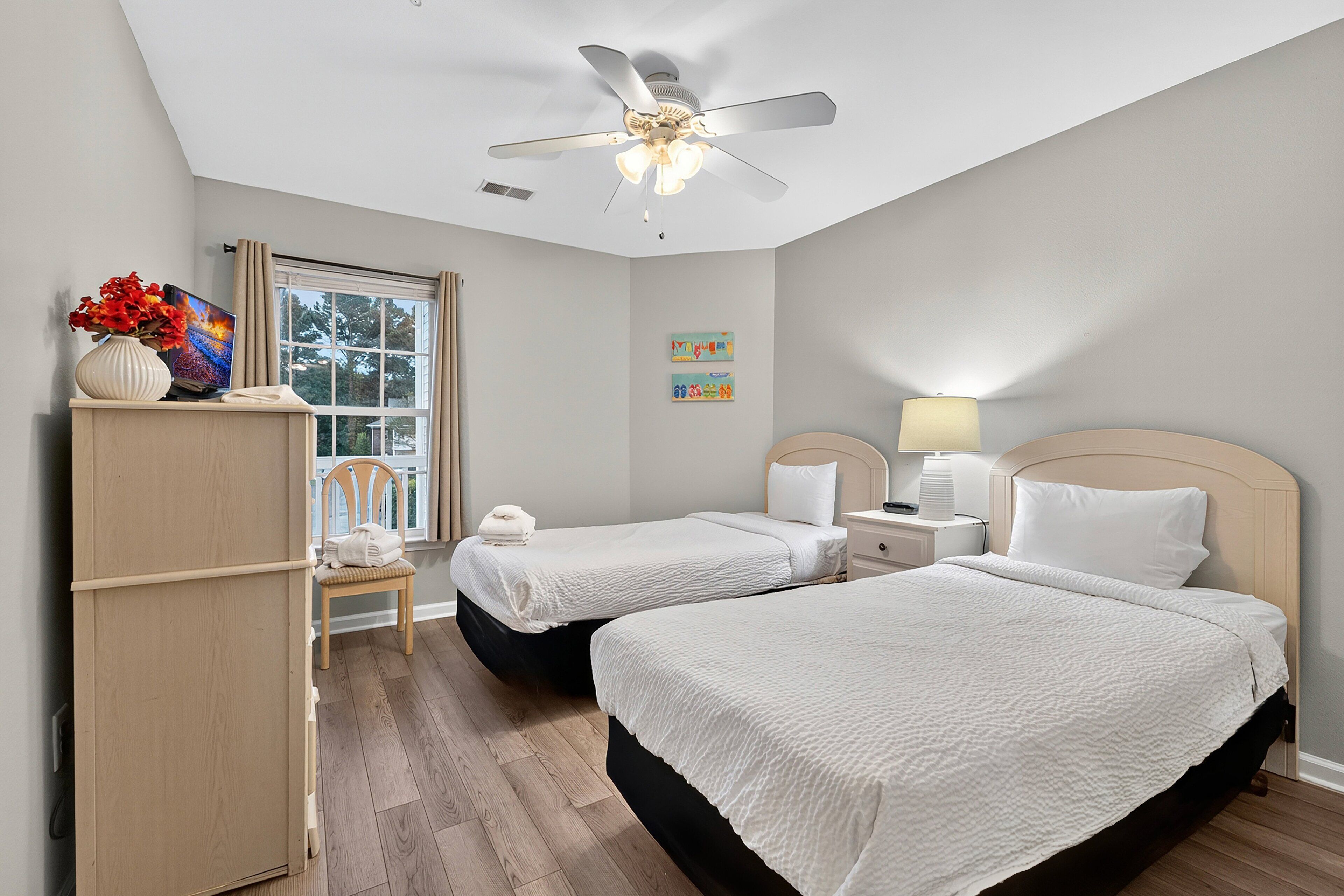 A bedroom with two beds, a ceiling fan, a window, and a nightstand with a lamp.