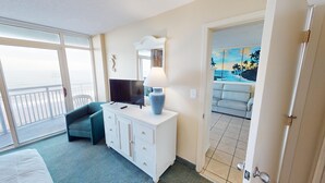Una camera d'albergo con balcone, TV, lampada, divano e un quadro raffigurante una spiaggia.