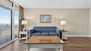 Uma sala de estar com um sofá azul, duas luminárias, um quadro na parede e uma janela grande com cortinas.