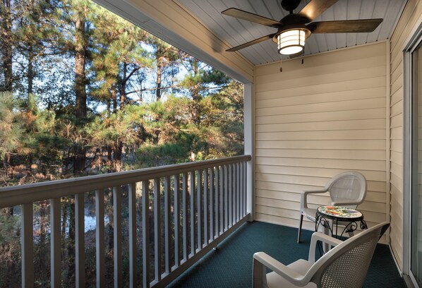 A covered porch with a ceiling fan, a table with a chair, and a view of a wooded area. - Fairways 26E (Myrtle Beach)