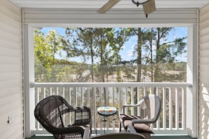 A covered porch with wicker chairs and a small table, overlooking a natural landscape with trees and a clear sky. - Fairways 26E (Myrtle Beach)
