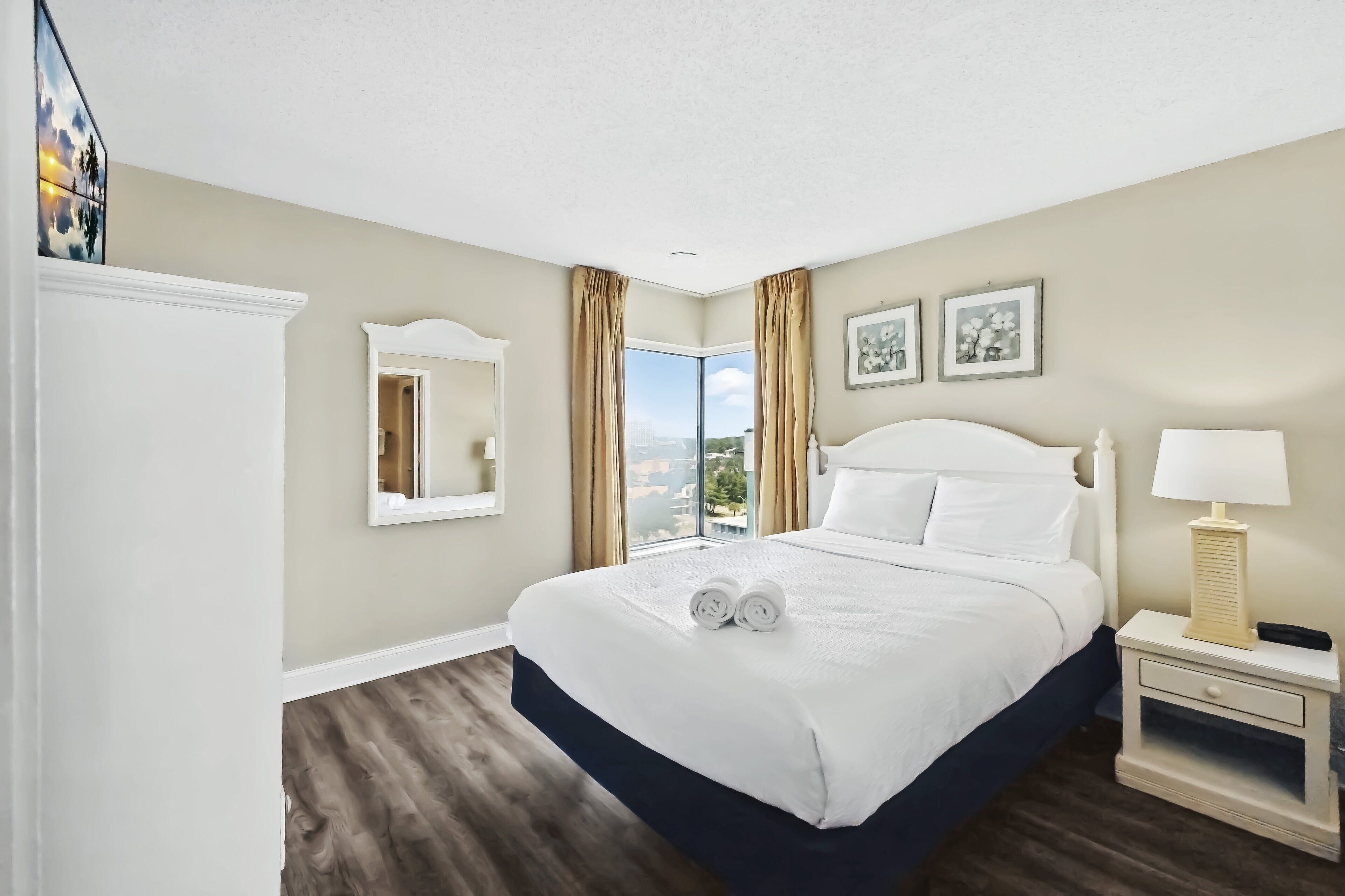 A hotel room with a large bed, a nightstand with a lamp, a mirror, and a view of the city through a large window.
