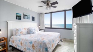 Een slaapkamer met een bed, een plafondventilator, een televisie, een raam met uitzicht op zee en een boekenplank.