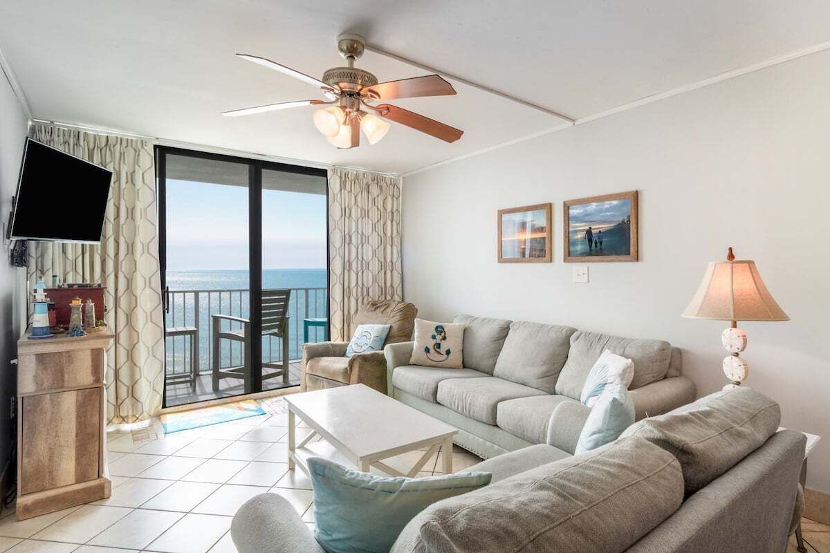 A modern living room with a sofa, coffee table, and a TV. There is a balcony with a view of the sea.