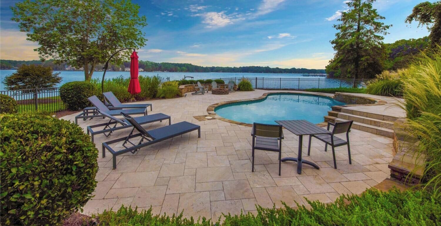 A swimming pool with lounge chairs and a table, surrounded by greenery and a view of a lake.