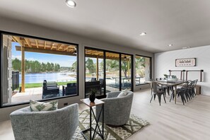 A spacious room with large windows overlooking a lake, a dining area with a wooden table and chairs, and a cozy seating area with grey upholstered chairs. - Salience Path (Mooresville)
