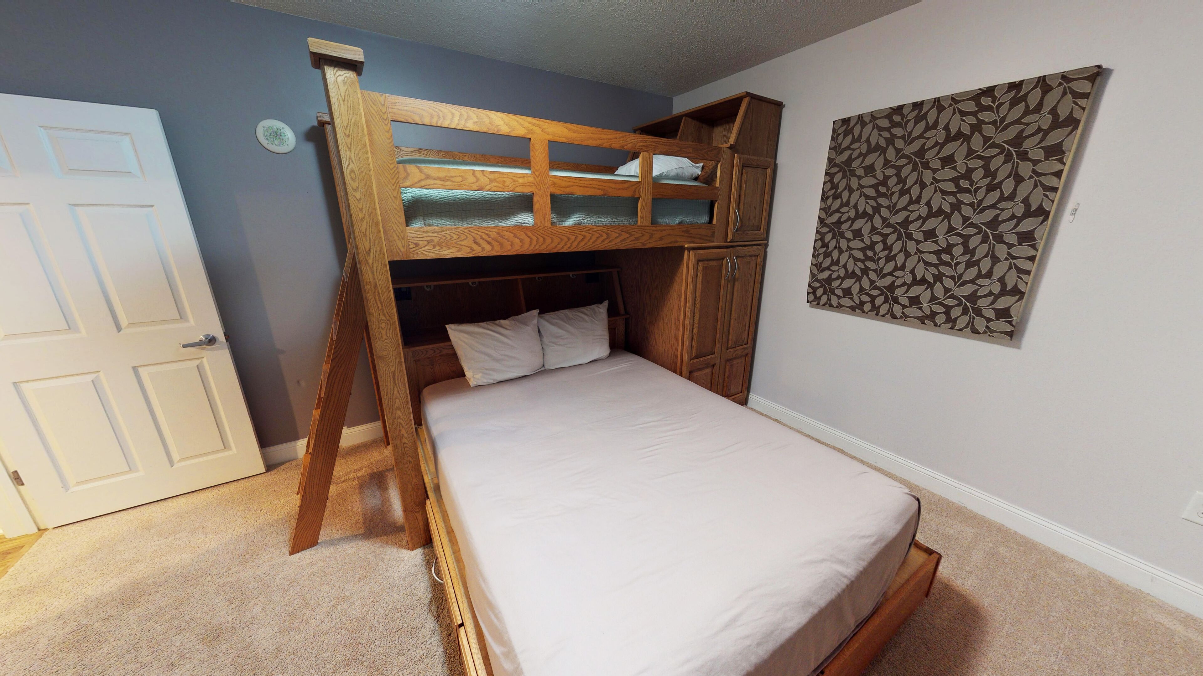 A bunk bed with a wooden frame and a lower bunk, a white door, and a wall with a leaf-patterned artwork.