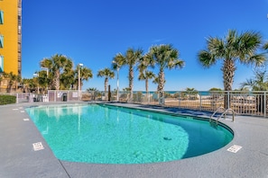 A swimming pool with clear blue water, surrounded by palm trees and a white fence. - Ocean Blue 1002 (Myrtle Beach)