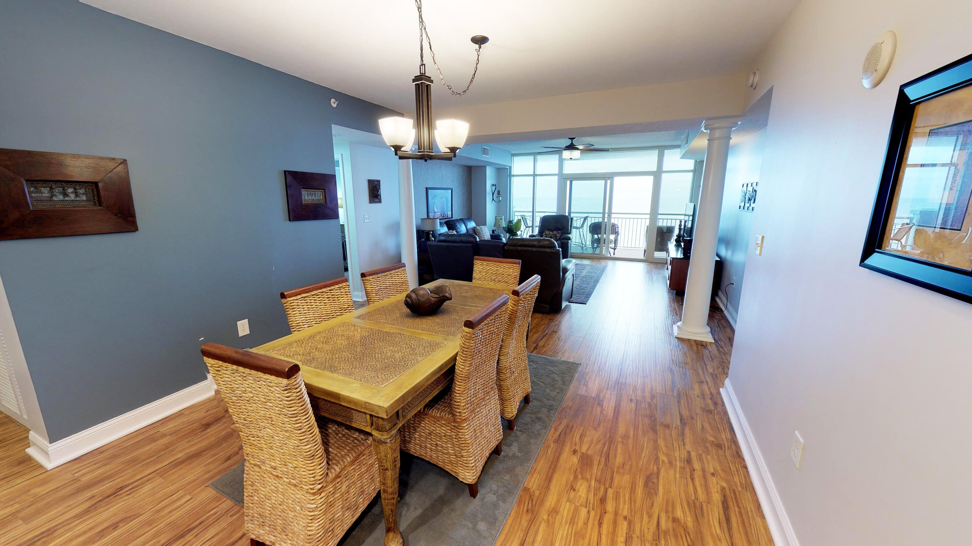 A dining area with a wooden table and wicker chairs, a chandelier, and a view to a living room with a sofa and chairs.
