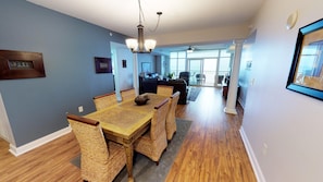 A dining area with a wooden table and wicker chairs, a chandelier, and a view to a living room with a sofa and chairs. - Ocean Blue 1002 (Myrtle Beach)
