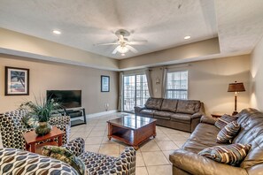 A spacious living room with a ceiling fan, a large window, and a brown leather sofa. - Myrtle Beach Villas 205 A (Myrtle Beach)