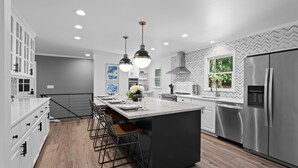 Une cuisine moderne dotée d’un îlot central, d’appareils électroménagers en acier inoxydable, de luminaires suspendus et d’un dosseret orné d’un motif en chevrons.