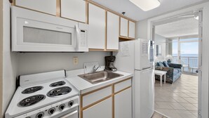A compact kitchen with a stove, microwave, sink, and refrigerator. A coffee maker is on the counter, and a sofa is visible in the adjacent living area. - Atlantica 903 (Myrtle Beach)