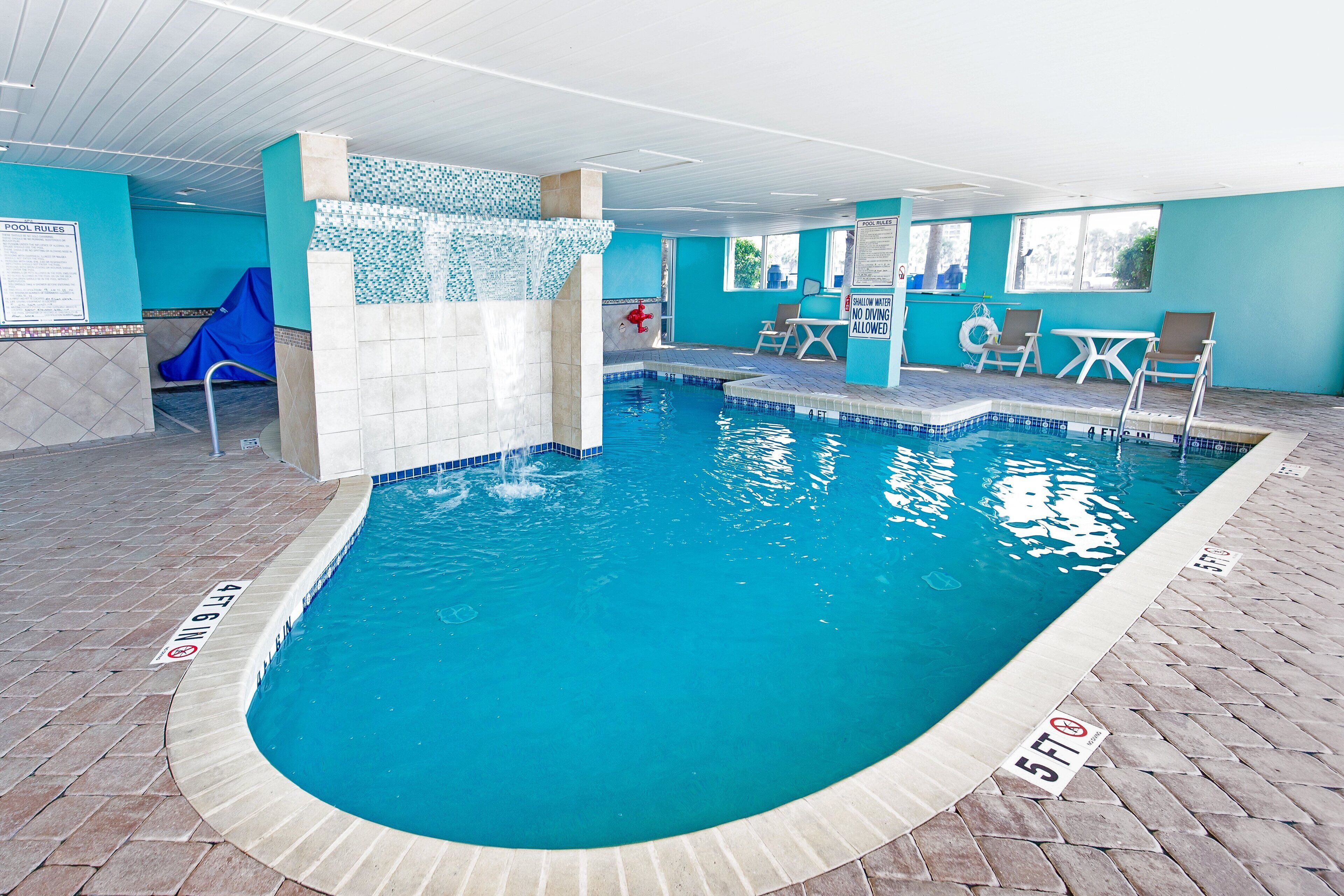 Une piscine intérieure avec un sol carrelé, un mur bleu et un espace aménagé au bord de la piscine, comprenant des chaises et une table.