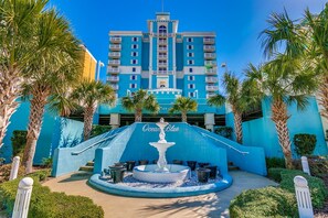 Ein Hotel Eingang mit einer Fontäne, Palmen und einem blauen Gebäude mit Balkonen.