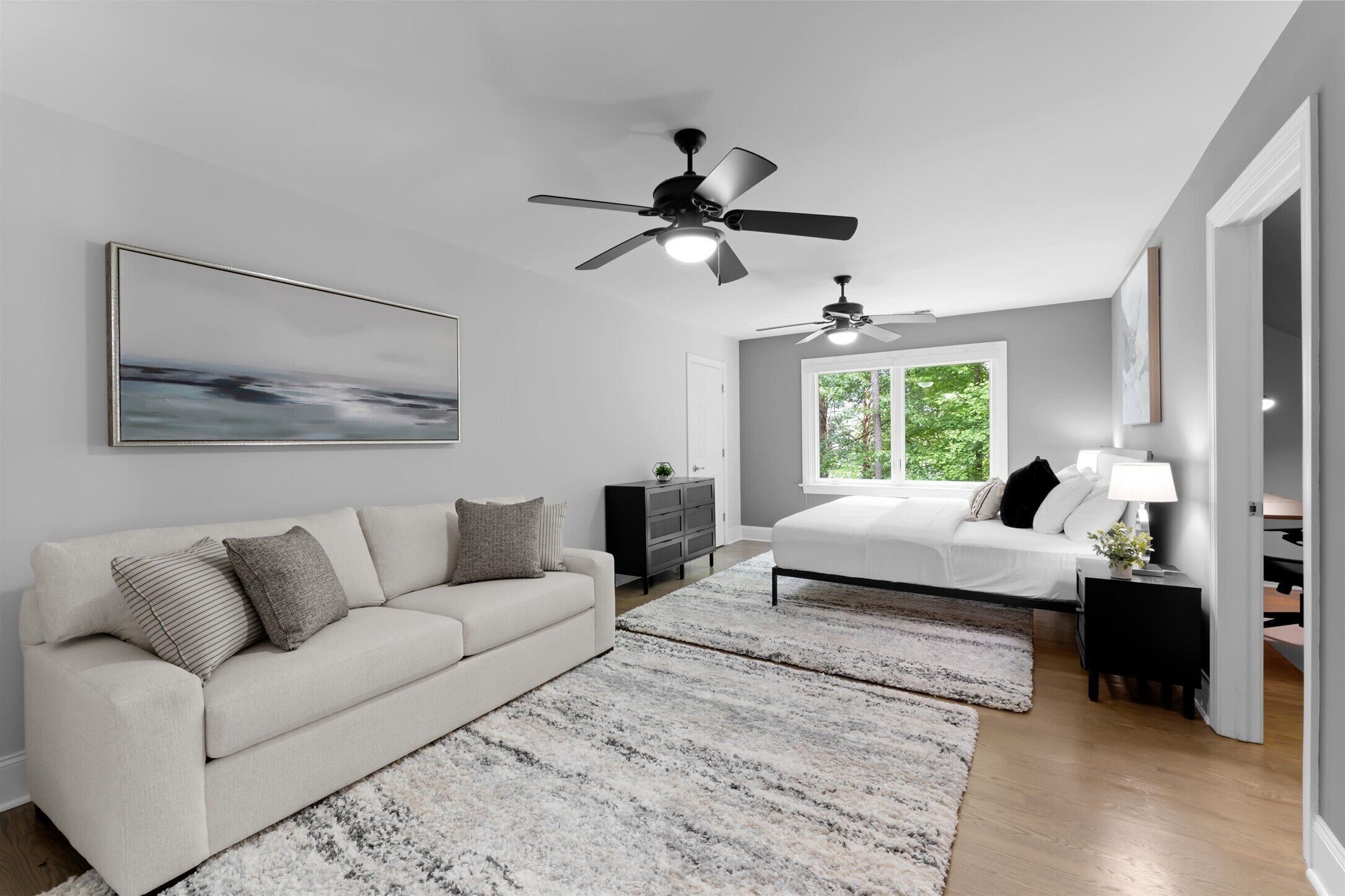 A modern bedroom with a bed, a sofa, a ceiling fan, and a window with a view of greenery.