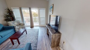 Un salon avec un canapé bleu, une table basse en bois, un téléviseur posé sur un meuble en bois et une grande fenêtre avec des rideaux.