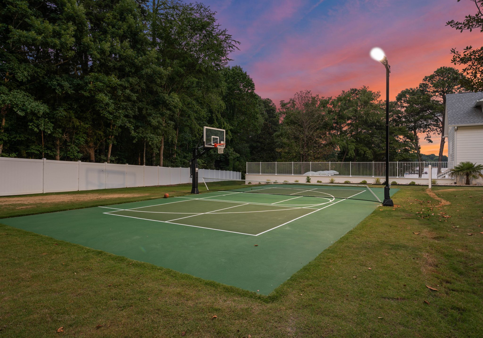 Un terrain de basket en plein air, doté d’une surface verte, entouré d’une clôture blanche et d’arbres.