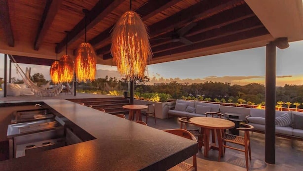 Uma área externa com mesas e cadeiras de madeira, um bar e luminárias pendentes.