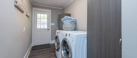 A laundry room with two front-loading washing machines, a basket on top of one, and a door leading to another room.