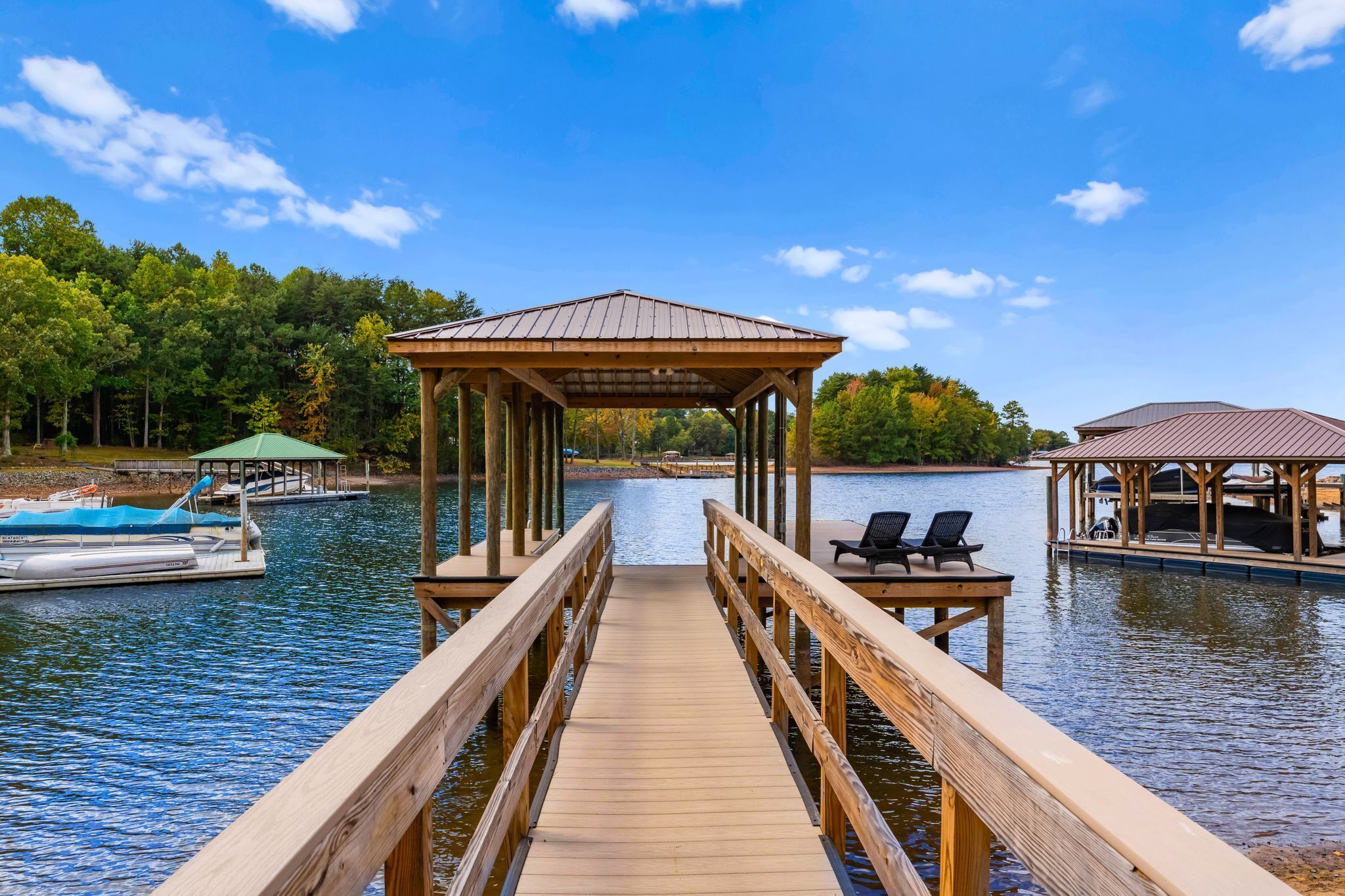 A wooden pier leading to a gazebo and a covered seating area by a calm lake with boats and trees in the background.