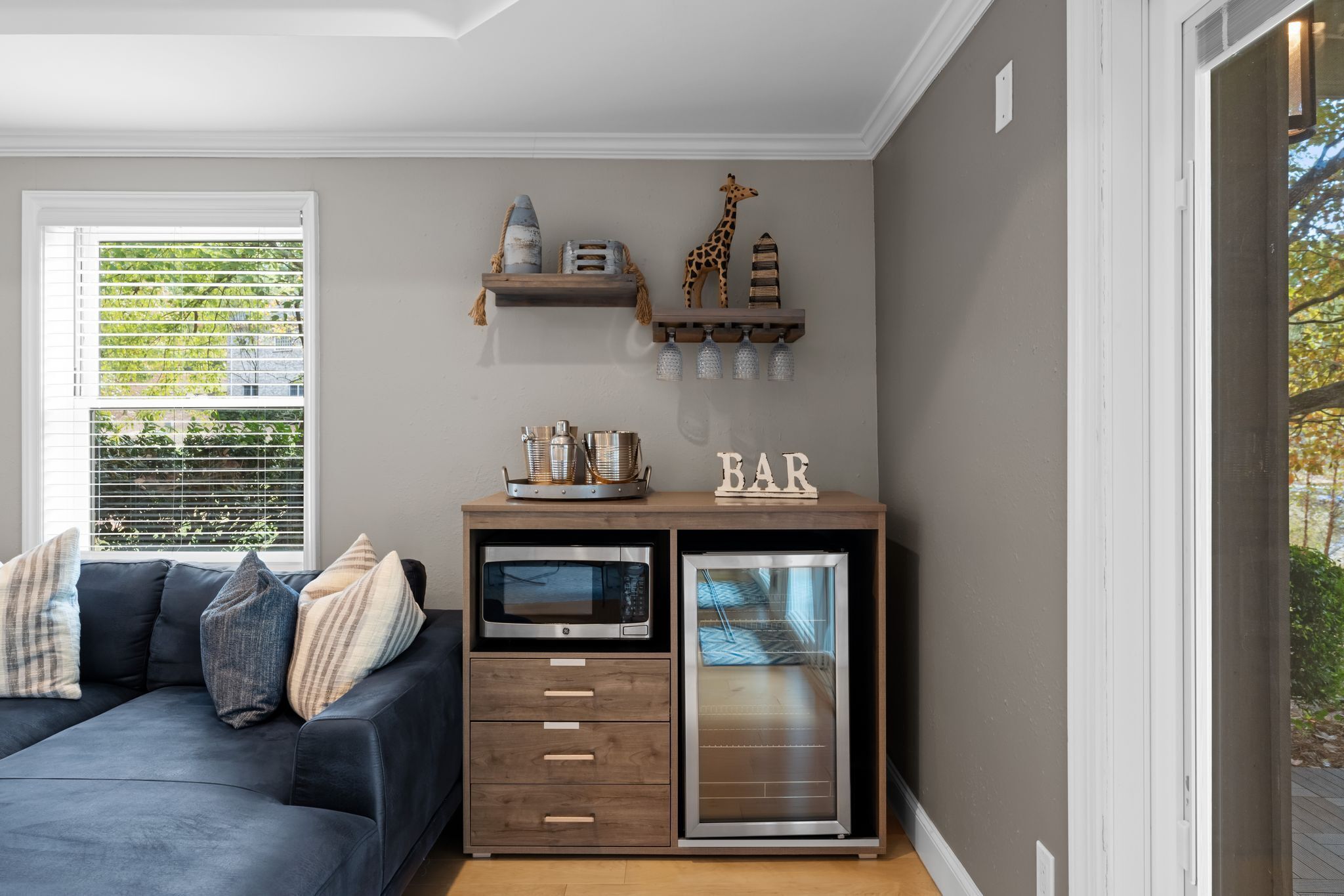 A modern living room with a dark sofa, a built-in microwave, and a mini-fridge. There are decorative shelves above the fridge and a window with blinds.
