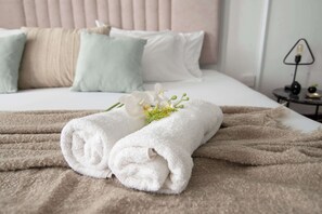 En pænt redt seng med sammenrullet hvide håndklæder, et beige tæppe og en lille buket hvide blomster.