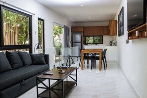 A modern living room with a sofa, coffee table, and dining area with chairs. There is a kitchen with wooden cabinets and a large TV. - Escapada de lujo en Playacar (Playa del Carmen)