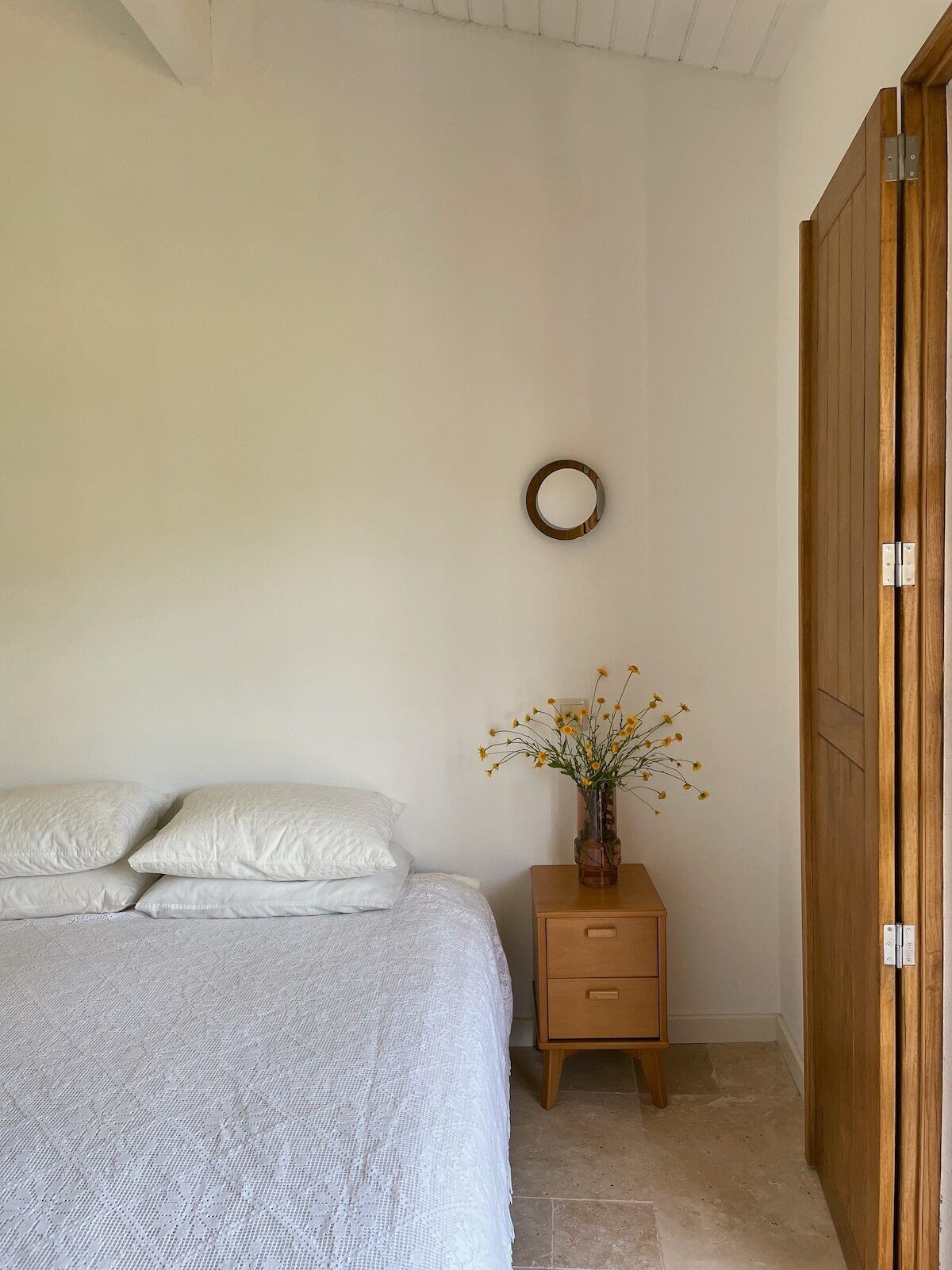 A bedroom with a bed, pillows, a nightstand with a vase of flowers, and a wooden wardrobe.