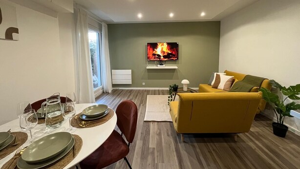 Ein modernes Wohnzimmer mit einem gelben Sofa, einem Essbereich mit einem runden Tisch und einem Fernseher, der ein Feuer zeigt.