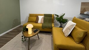 Ein Wohnzimmer mit einem senffarbenen Sofa, einem kleinen Beistelltisch mit Lampe und einem Blumentopf.