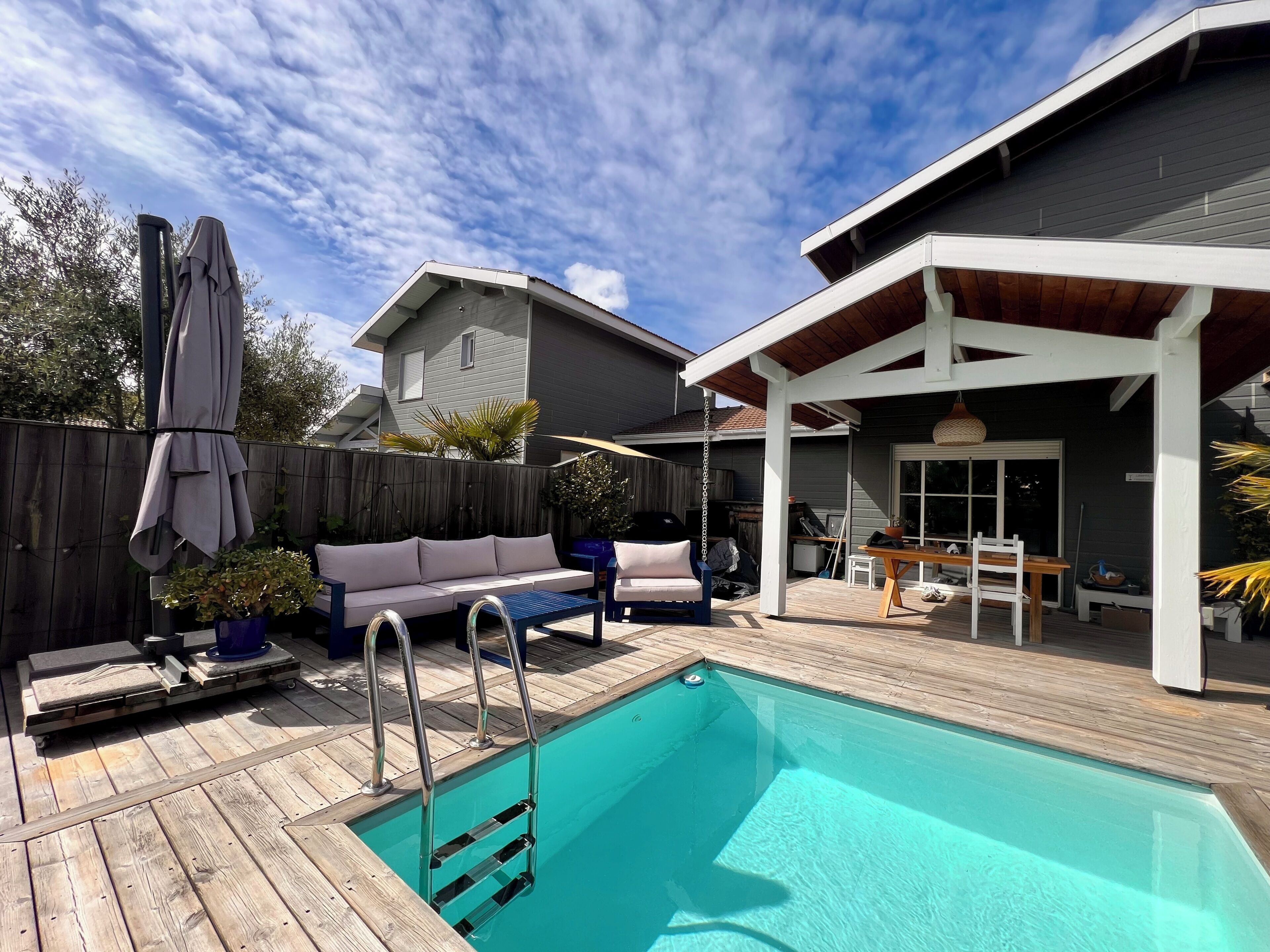 A backyard with a swimming pool, wooden deck, outdoor furniture, and a covered patio area.