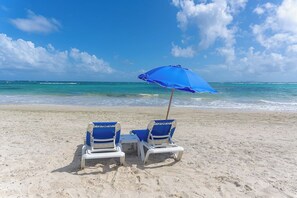 Zwei Strandstühle mit einem blauen Sonnenschirm am Sandstrand.