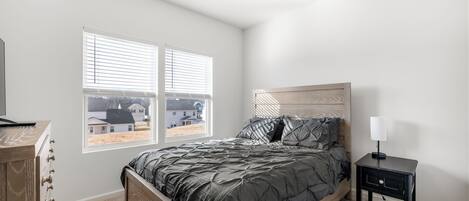Un dormitorio con cama, mesita de noche, cómoda y dos ventanas con persianas.