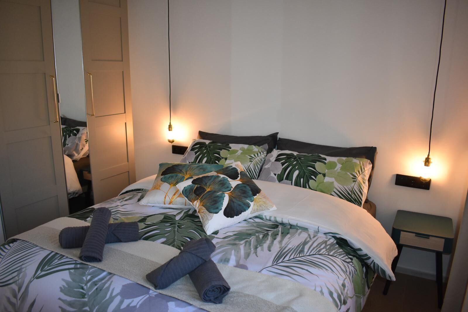 A neatly made bed with tropical leaf-patterned bedding, two bedside tables with wall-mounted lamps, and a mirror on the left wall.